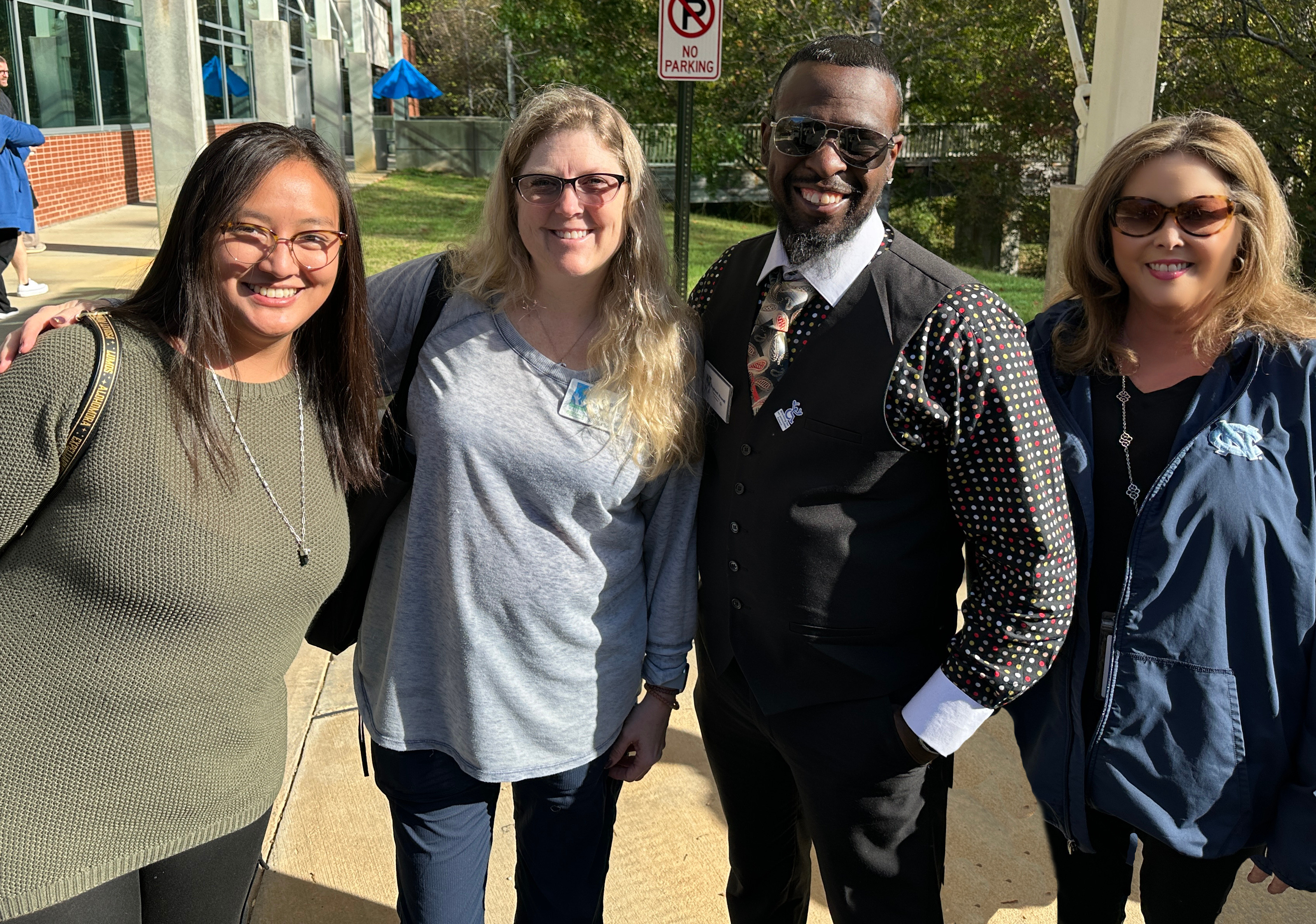 Three women and a man smiling and standing in a parking lot. 