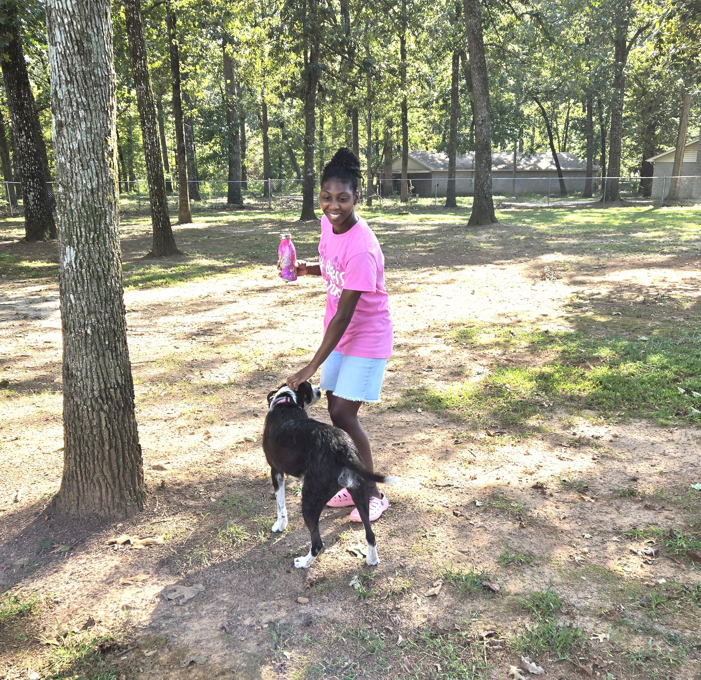 A girl in a pink shirt and denim shorts is petting a dog and holding a water bottle. 