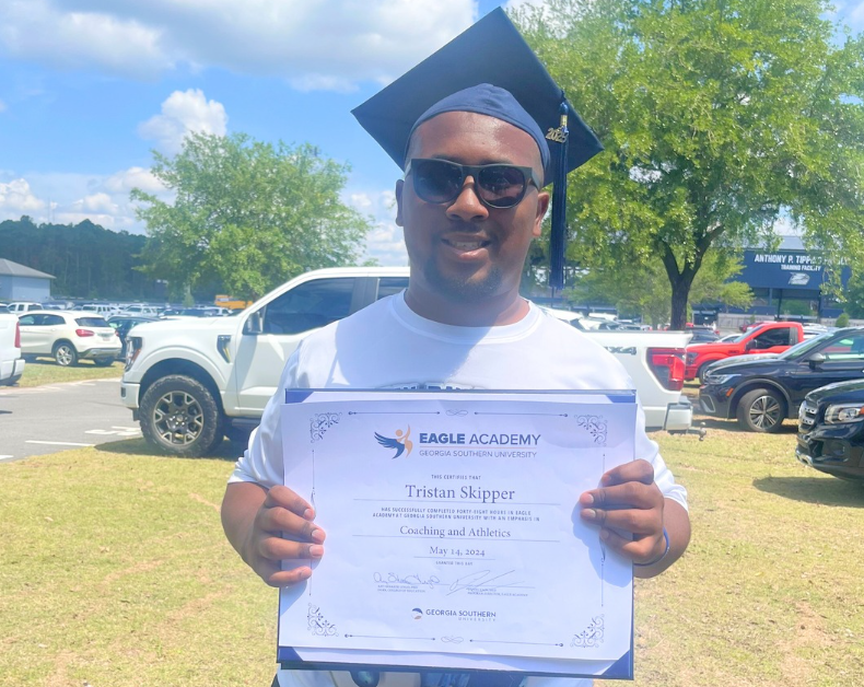 A male with a graduation cap wearing black sunglasses and holding up a certificate. 