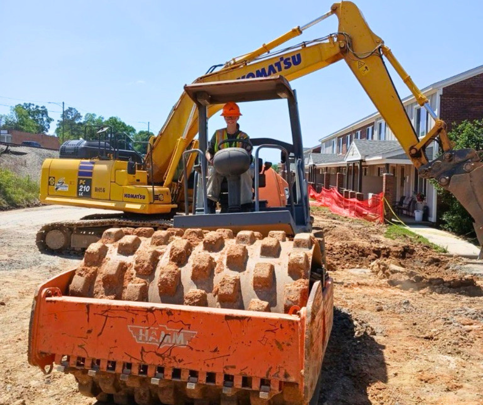 A person in an orange hard hat and safety vest operates a large orange soil compactor with a textured roller in front of a yellow excavator at a construction site.