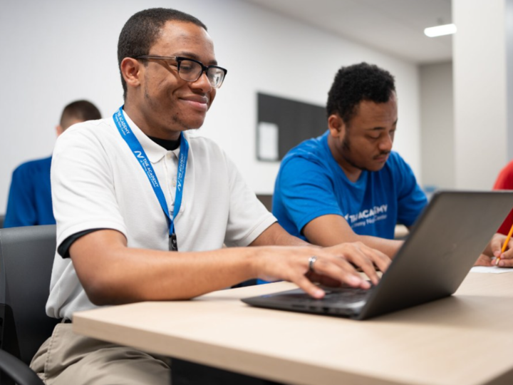 A student in a classroom smiling while looking at his laptop. 