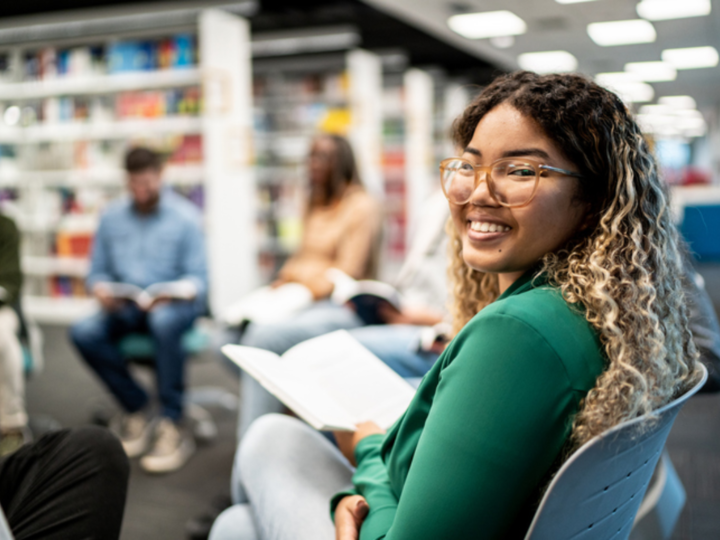 A student that is smiling with a book in her hand and in the background with  other students around .