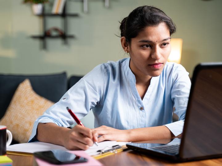 A young teen writing down information on a notepad while looking at her laptop.
