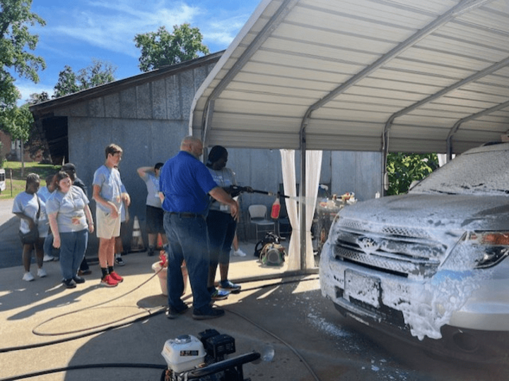 A student holding a water hose and spraying water on the car. 