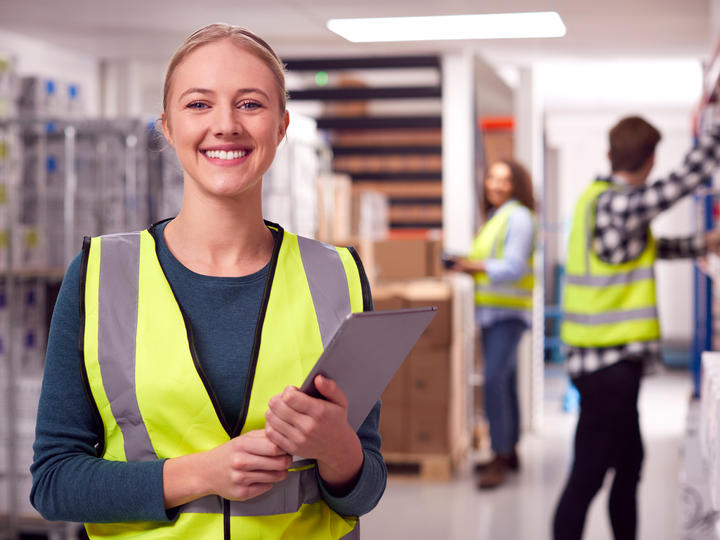 A female warehouse worker who is smiling and holding a notepad. 