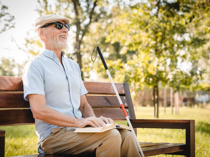 An older blind man wearing a white hat and black sunglasses. 
