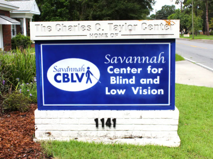There is a blue and white sign outside the Savannah Center for Blind and Low Vision. The sign reads " The Charles C. Taylor Center Home of the Savannah Center for Blind and Low Vision."