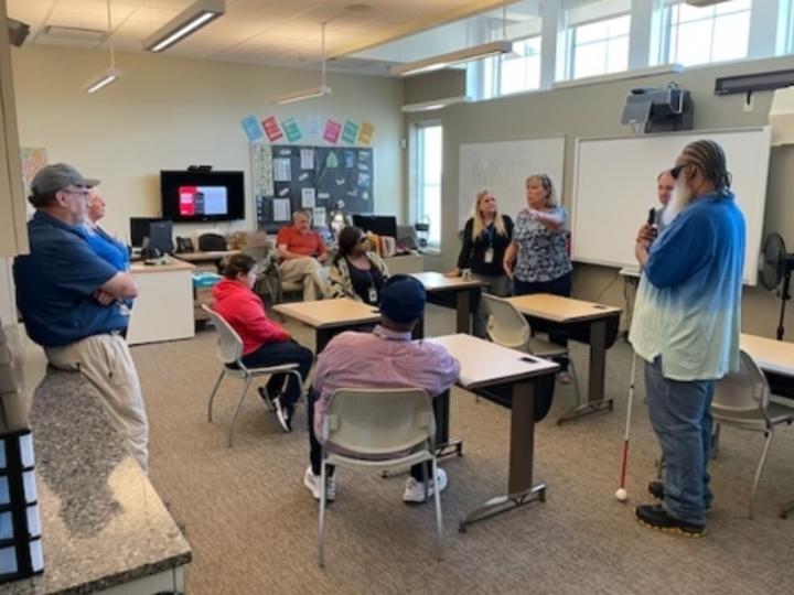 A group of people in a classroom listening to a woman speak in front of the classroom. 