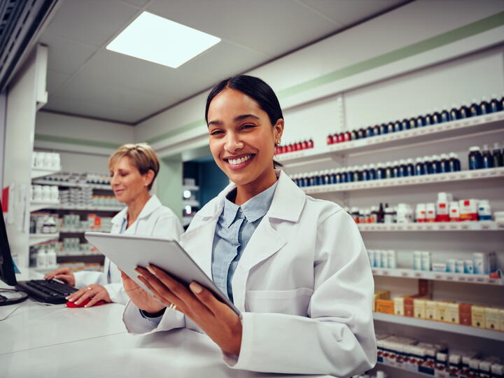 Two women that are Pharmacy Tech employees. One of the women is holding a tablet and smiling. 