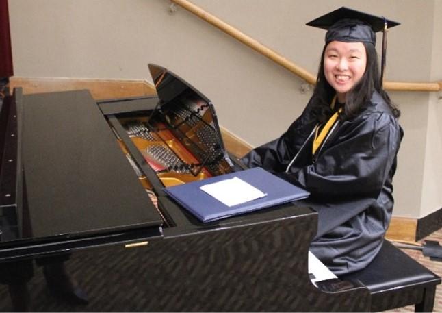 Anney Kim wearing a graduation gown and cap, and sitting at a piano.