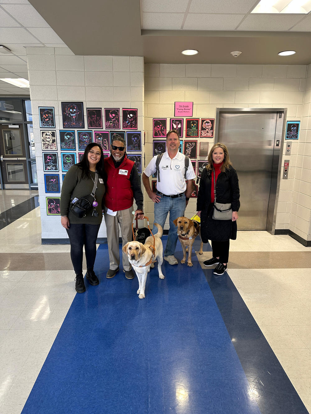 Two women and two men with service dogs standing in front of an elevator at a school. 