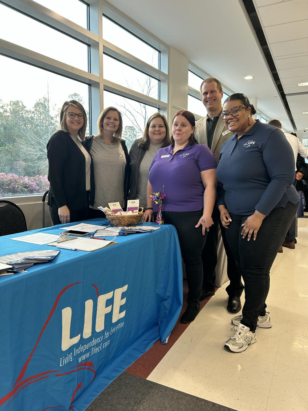 5 women and 1 male pose in front of a large window at an event