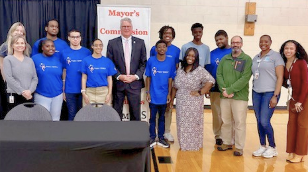 A group photo of students wearing blue shirts and the Mayor along with other county officials.