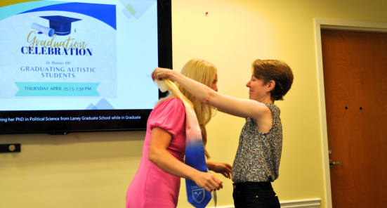 Two women face to face and one of the women is placing a graduation stole over a woman's head.  