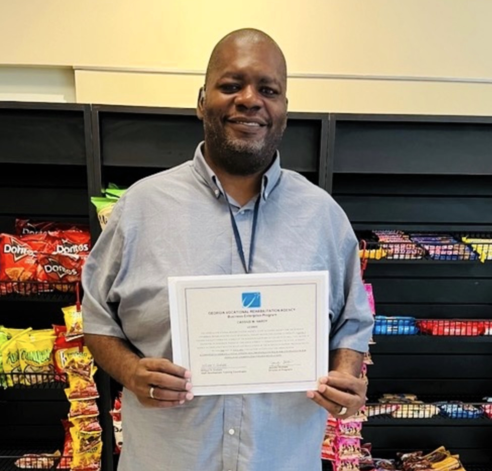 A man that is standing behind a black shelf filled with snacks holding up a certificate and smiling. 
