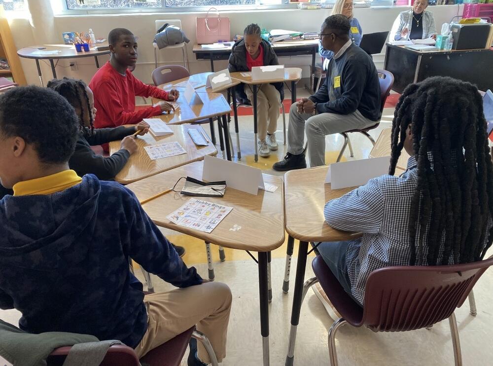 A group of students sits in a semi-circle with a facilitator in the middle.