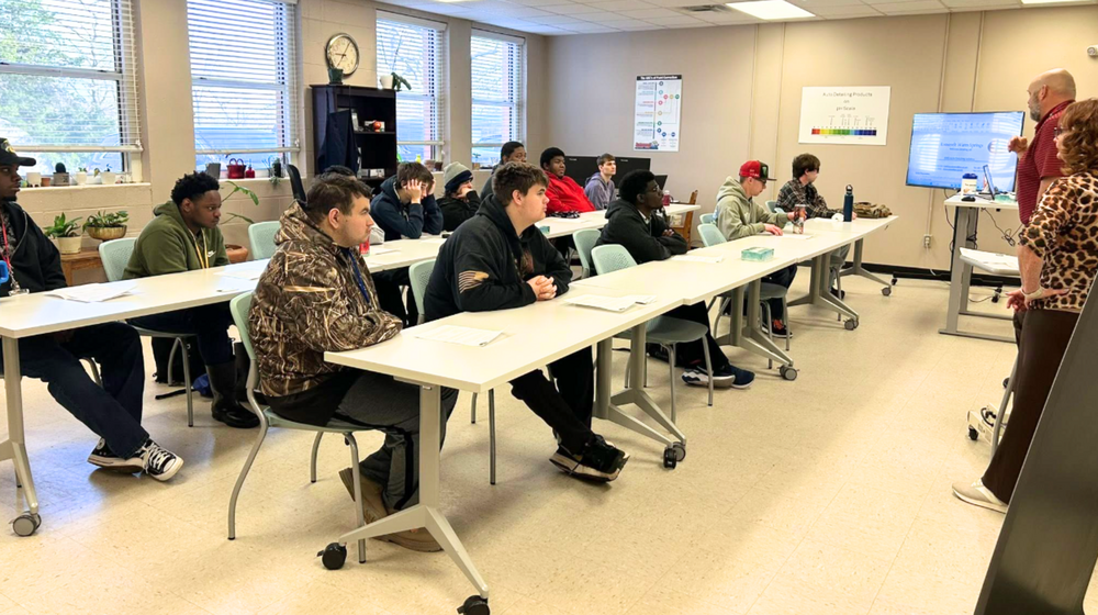Students in a classroom sitting at a table and two instructors are in front of the class talking. 