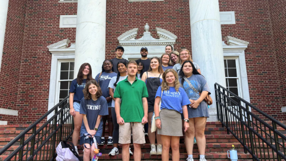 A group of students wearing blue Berry College shirts and one student is wearing a green shirt standing in front of a brick building. 