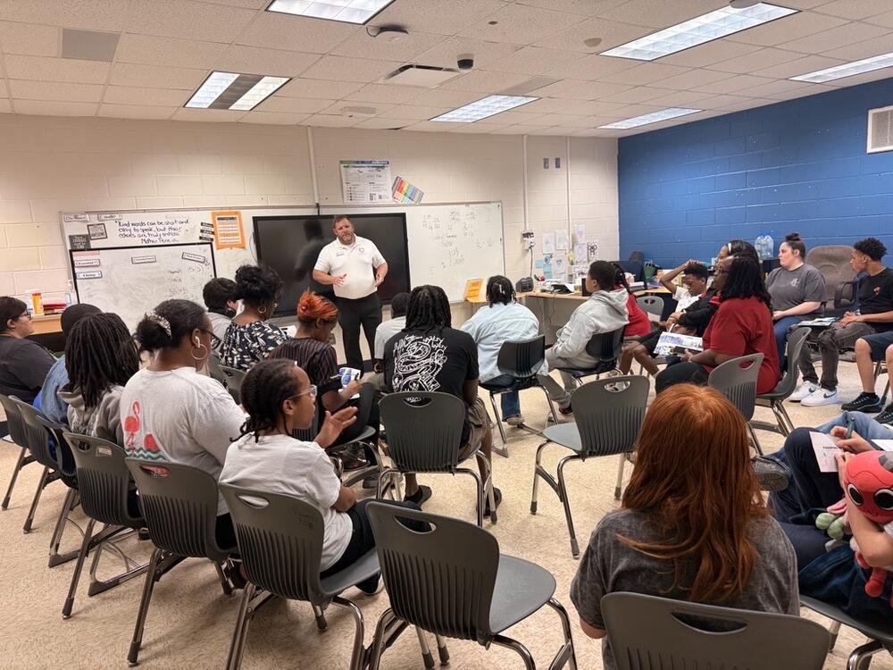 Students and adults sitting in a classroom, listening to a speaker