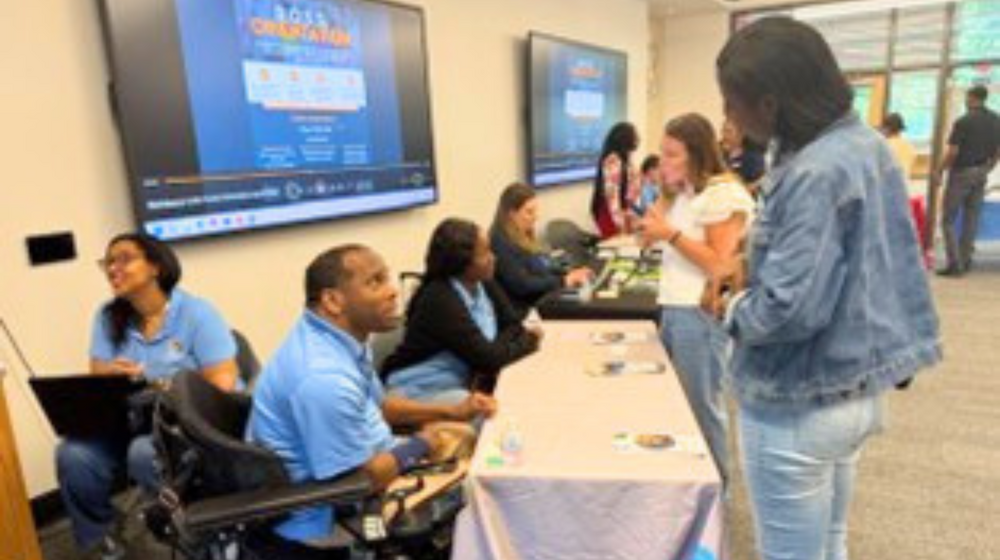 A man wearing a blue GVRA t-shirt talking to a woman wearing a jean jacket and blue jeans. 
