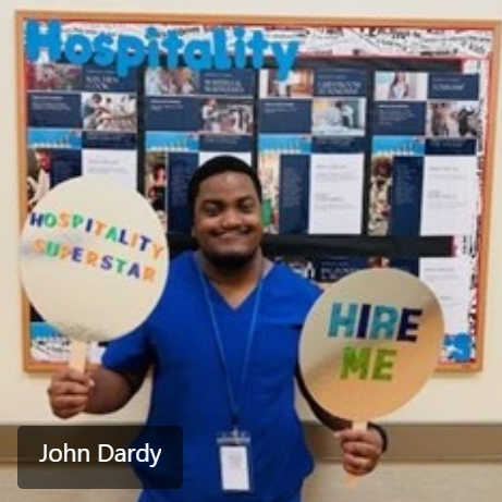 A man wearing a blue shirt and holding up signs that says Hire Me and Hospitality Superstar. 