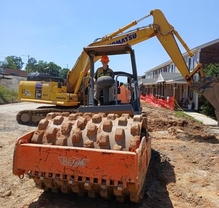 A person in an orange hard hat and a safety vest operates a large orange soil compactor with a textured drum, with a yellow excavator in the background on a construction site.