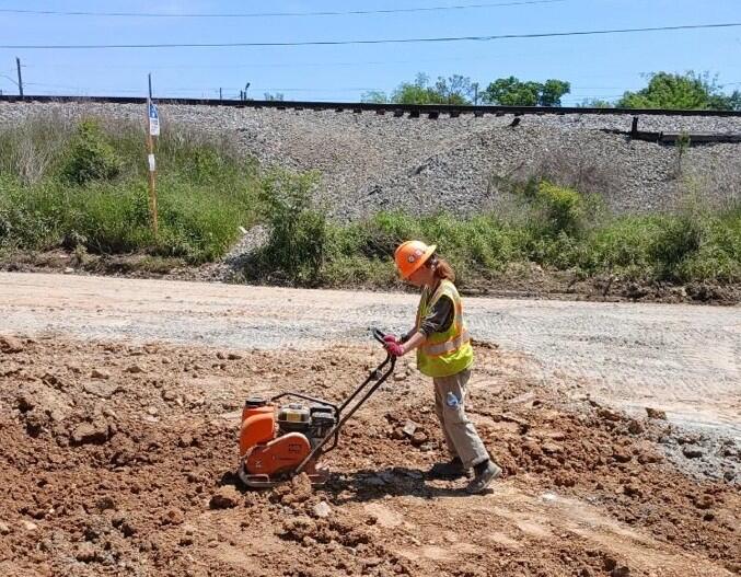A person in a hard hat and safety vest operates a plate compactor on a dirt construction site with railroad tracks in the background.