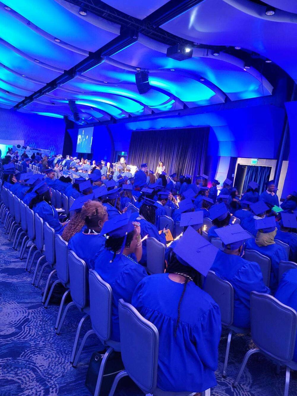 Graduates sitting down wearing blue graduation gown and cap watching the presenter speak. 