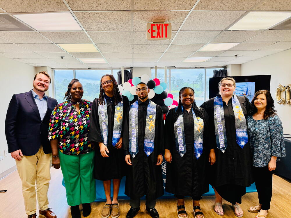 Seven people standing side by side and four of them are wearing graduation gown with a blue and black stole. 