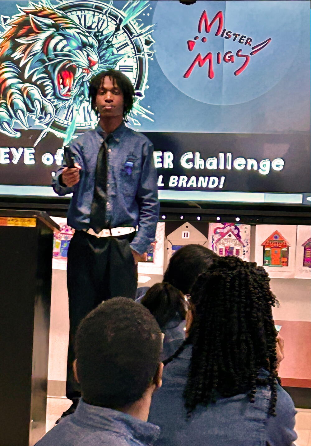 A young person stands at a podium in front of a screen displaying "Mister Mig's" logo and "EYE of... ER Challenge L BRAND!".