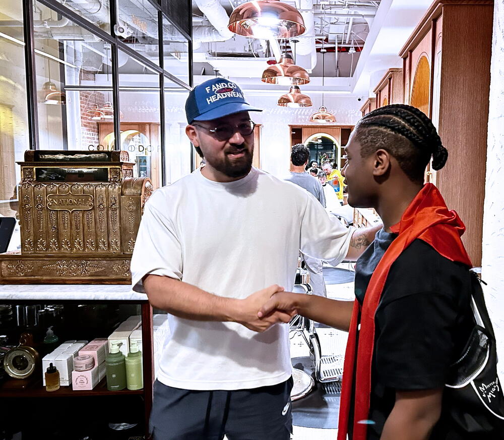 Two people shaking hands. One person wears a white t-shirt and a blue baseball cap. The other person wears a black t-shirt with an orange cloth draped over their shoulders and has braided hair.