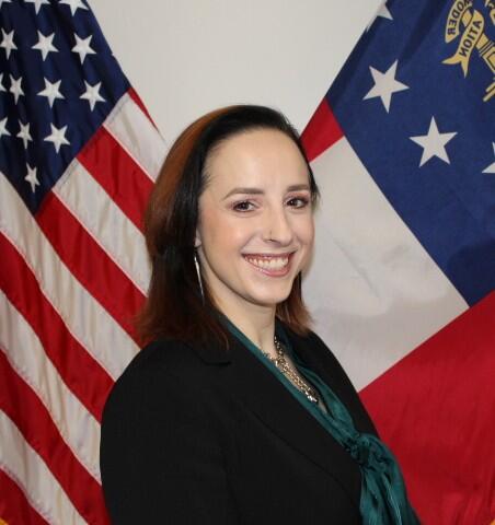 A woman in a black blazer and teal scarf smiles in front of the American flag and the Georgia state flag.