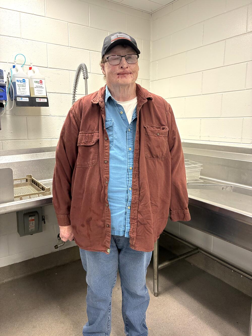 A person wearing glasses, a blue shirt, a reddish-brown jacket, and jeans stands in a room with sinks and tiled walls.