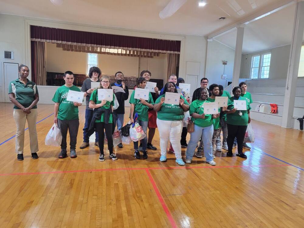 A group of students and Tree of Life staff wearing green shirts standing in front of a stage and holding up a document in their hand. 