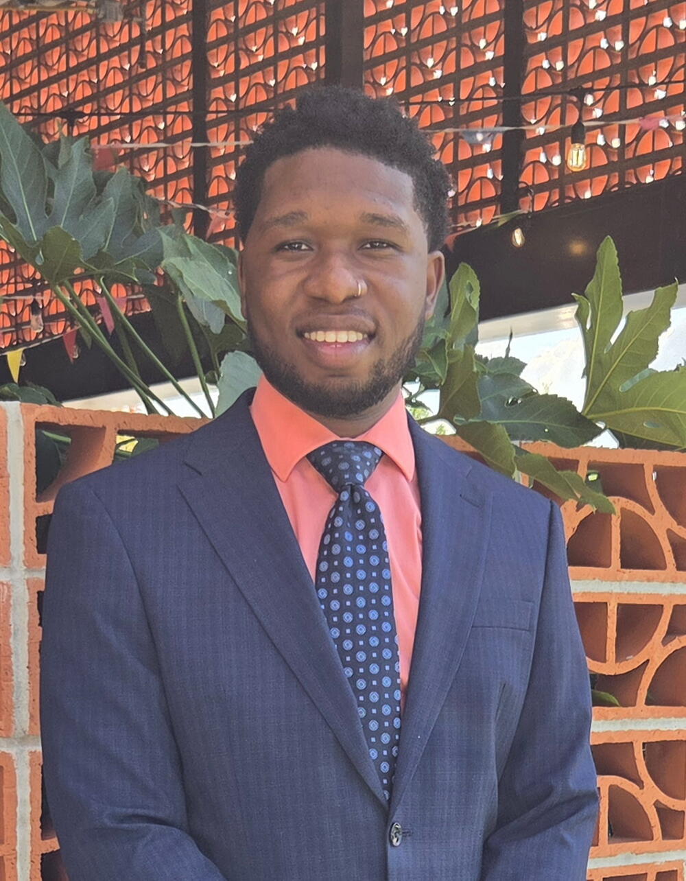 A man in a dark suit and coral dress shirt with a navy patterned tie smiles at the camera, standing outdoors in front of an orange brick wall and a lattice with warm lights and green leaves.