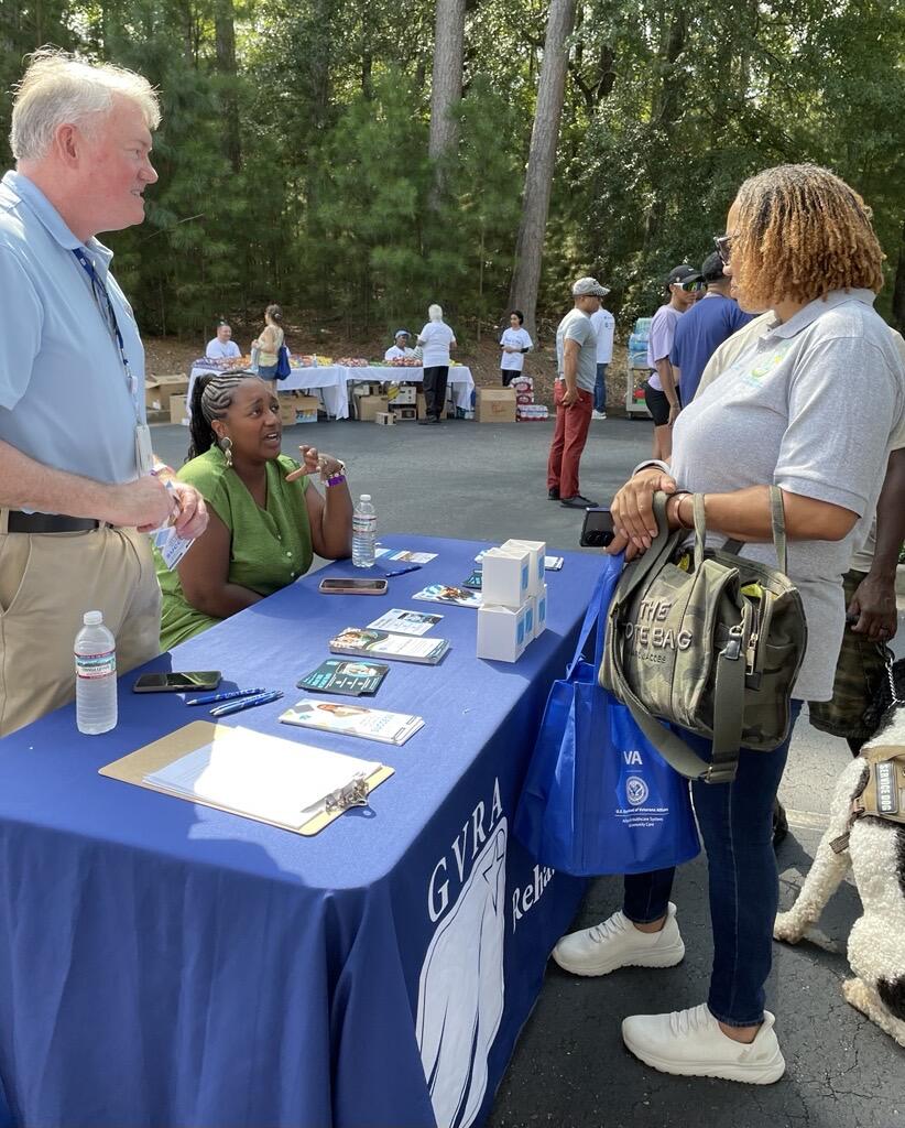 Outdoor community event at a blue table with brochures and card displays; several people stand and talk while others sit in the background under trees.
