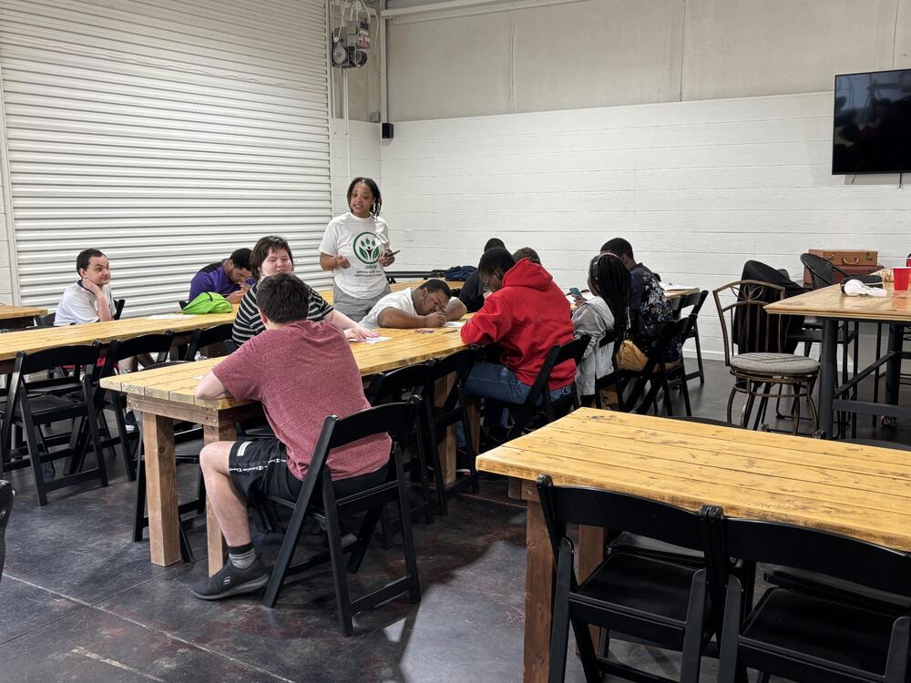 Students sitting down at the table and instructor is standing and talking to the students. 