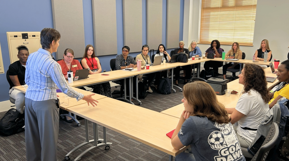 A group meeting around a long rectangular table in a classroom or conference room, with one person standing and gesturing while others sit and listen.