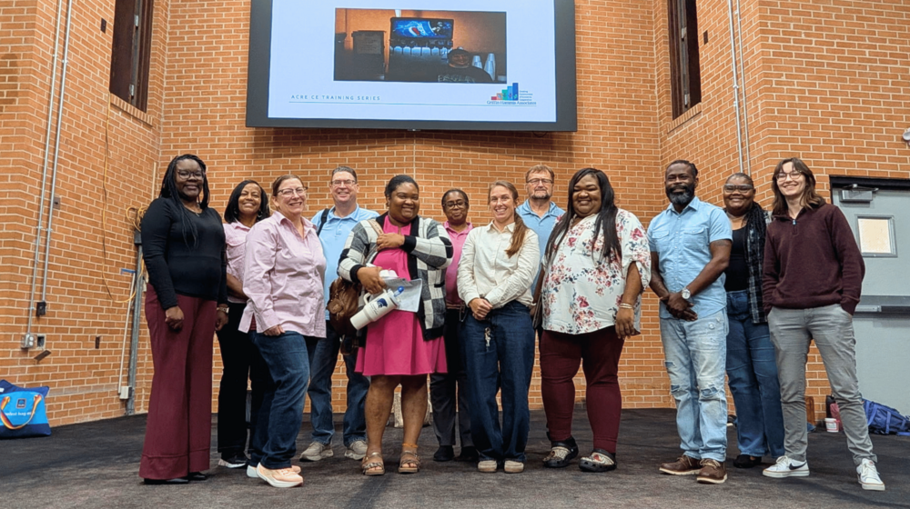 A group of people standing beside each other in front a large projector screen and brick wall. 