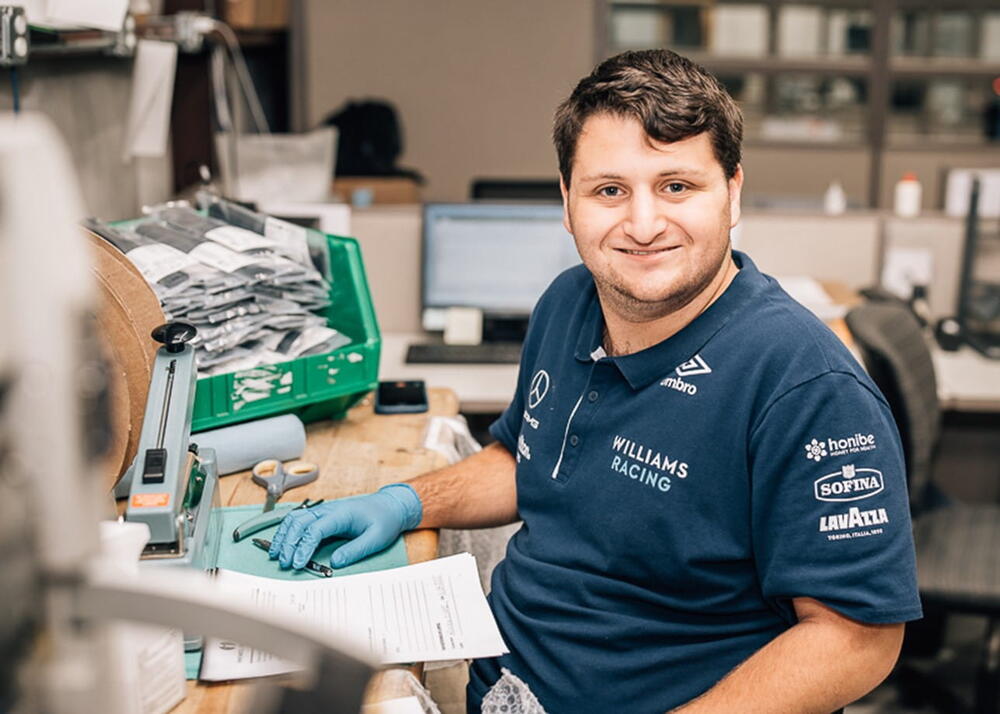 A technician in a blue work shirt sits at a desk in a workshop, wearing blue gloves and smiling while adjusting a green machine; papers and tools are scattered around.