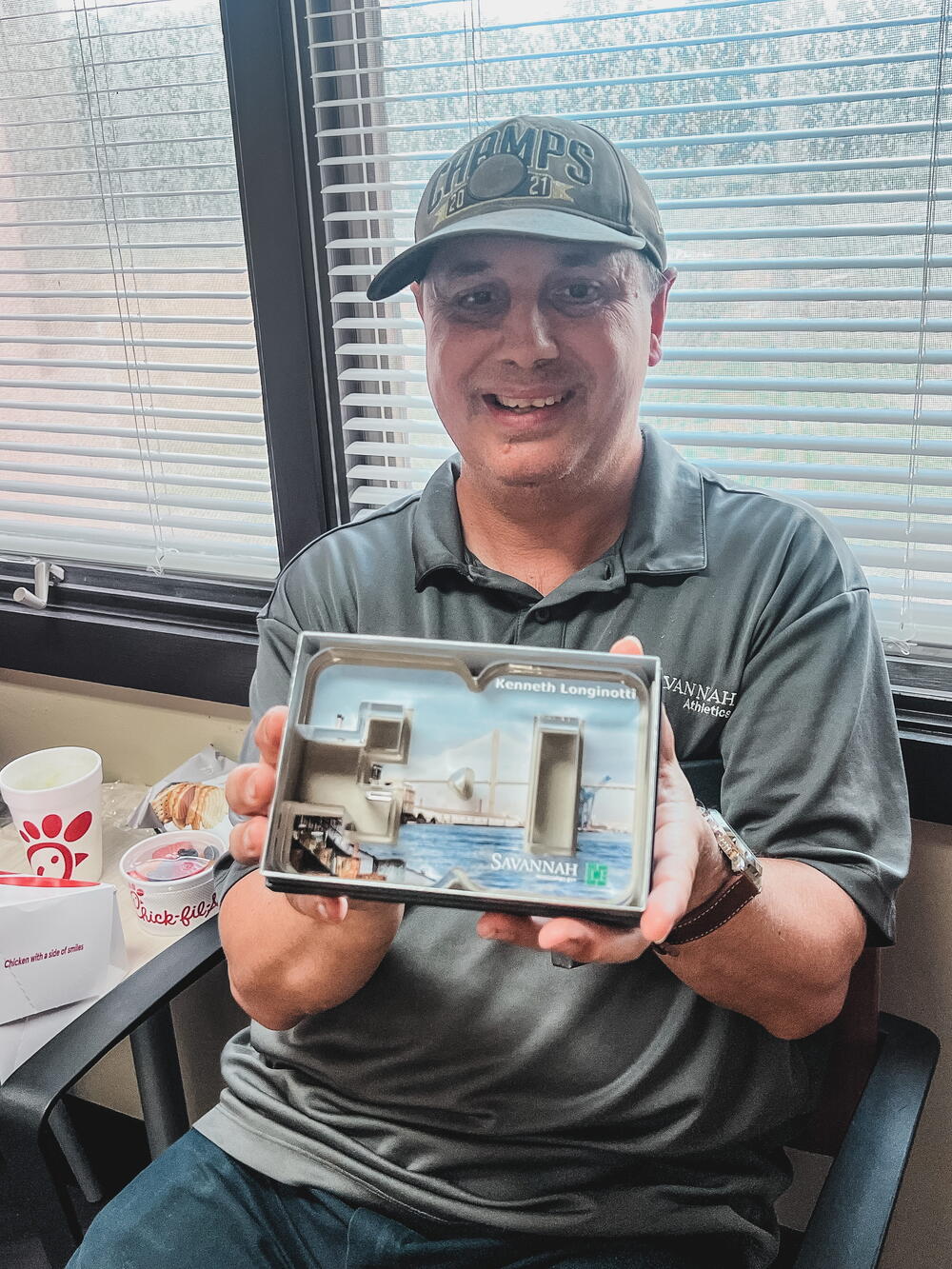 A man wearing a baseball cap sits at a table and smiles while holding a small framed architectural model labeled “Savannah.” Behind him are office blinds and a window.