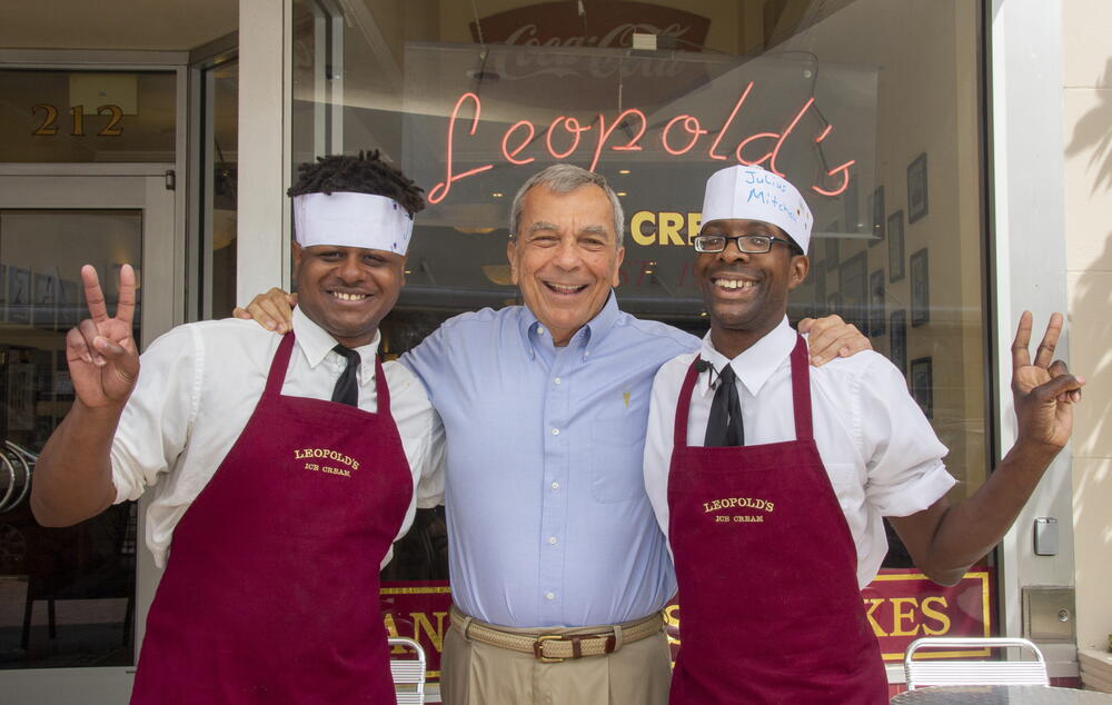 Two chefs in burgundy aprons and white headbands pose outside a restaurant entrance, smiling and making peace signs.