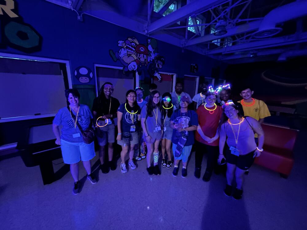 A group of people gathering in a dimly lit room with glow stick necklaces on. 