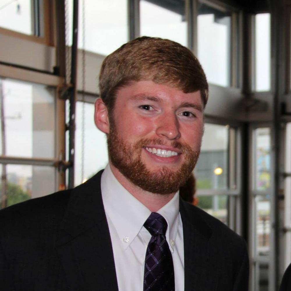 White male strawberry blonde hair with black suit jacket, white shirt, and black and blue tie