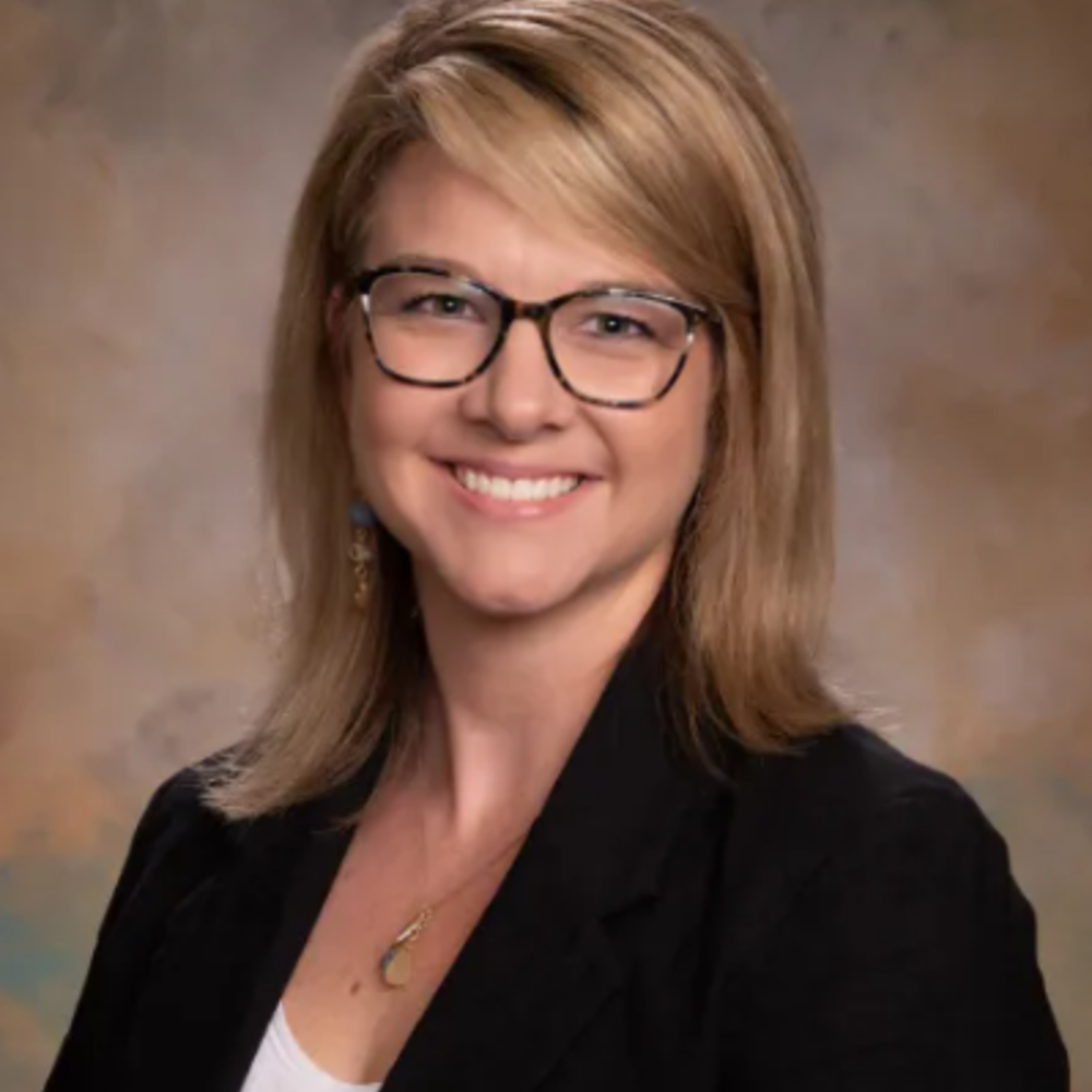 White woman with brownish blonde hair smiling, wearing glasses, a black blazer and white shirt