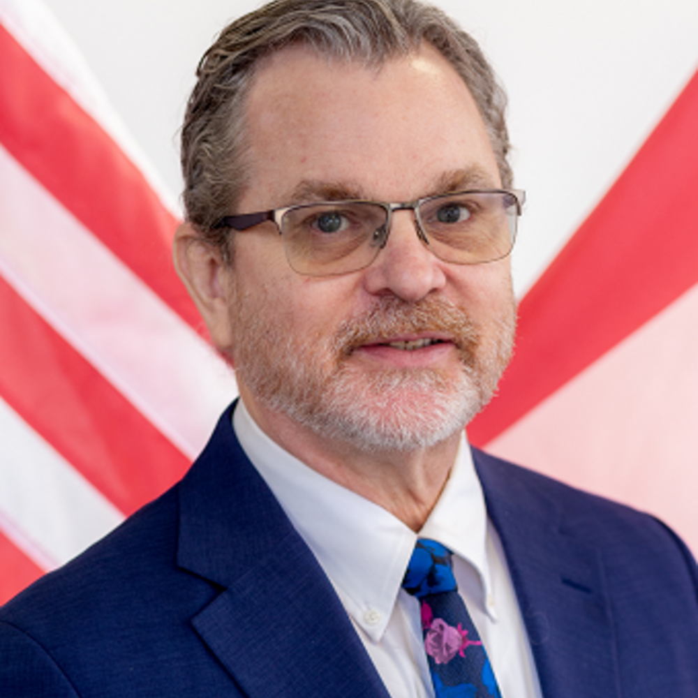 A photo headshot of Greg wearing a dark blue suit. 