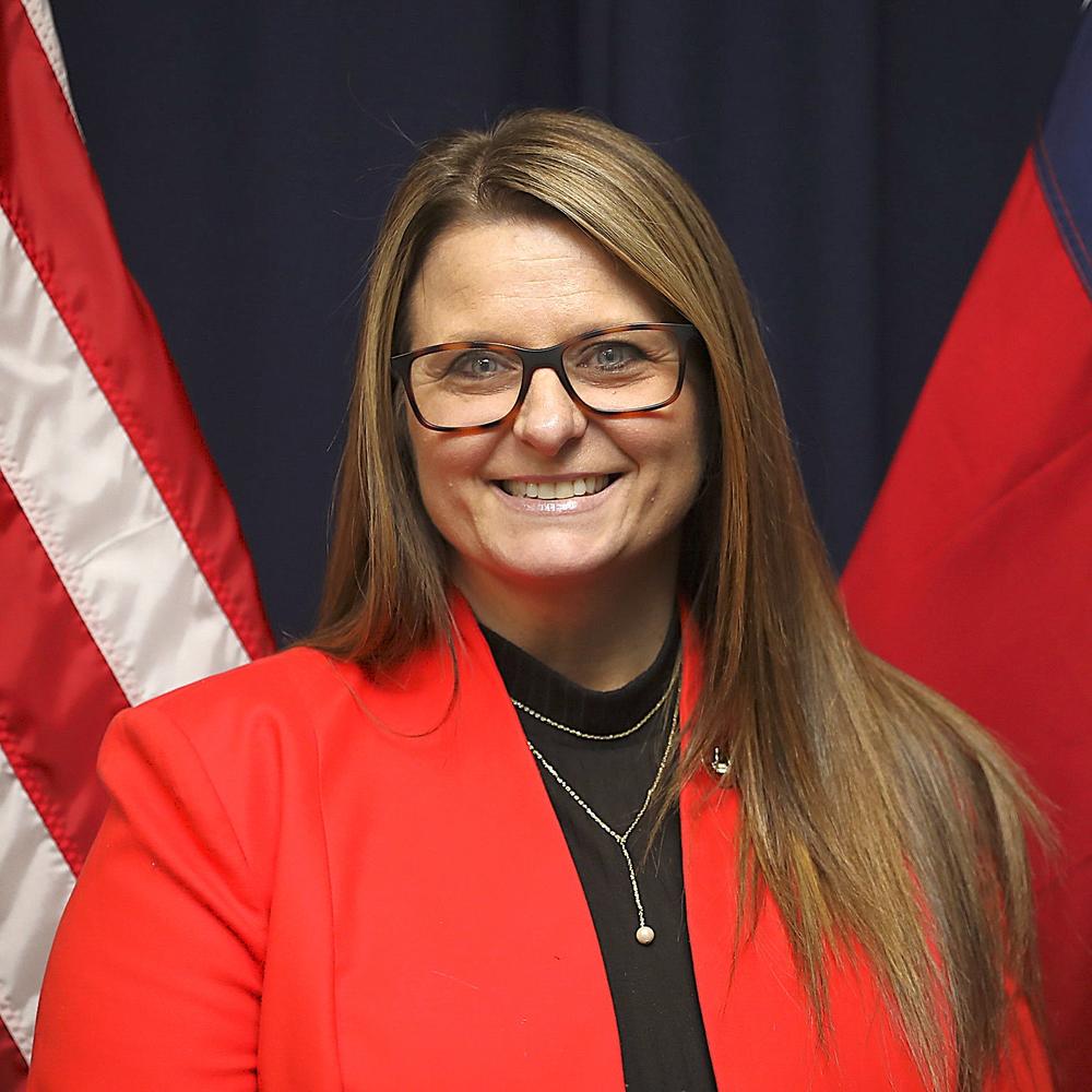 A headshot photo of a woman with a red blazer and that is wearing glasses.