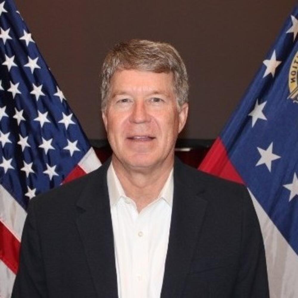 Male with brown graying hair wearing a white shirt with a black jacket