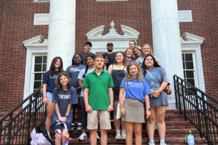 A group of students wearing blue Berry College shirts and one student is wearing a green shirt standing in front of a brick building. 