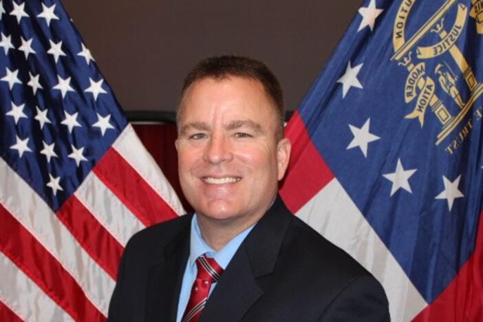A man wearing a black suit and a red a tie behind an American and state of Georgia flag. 